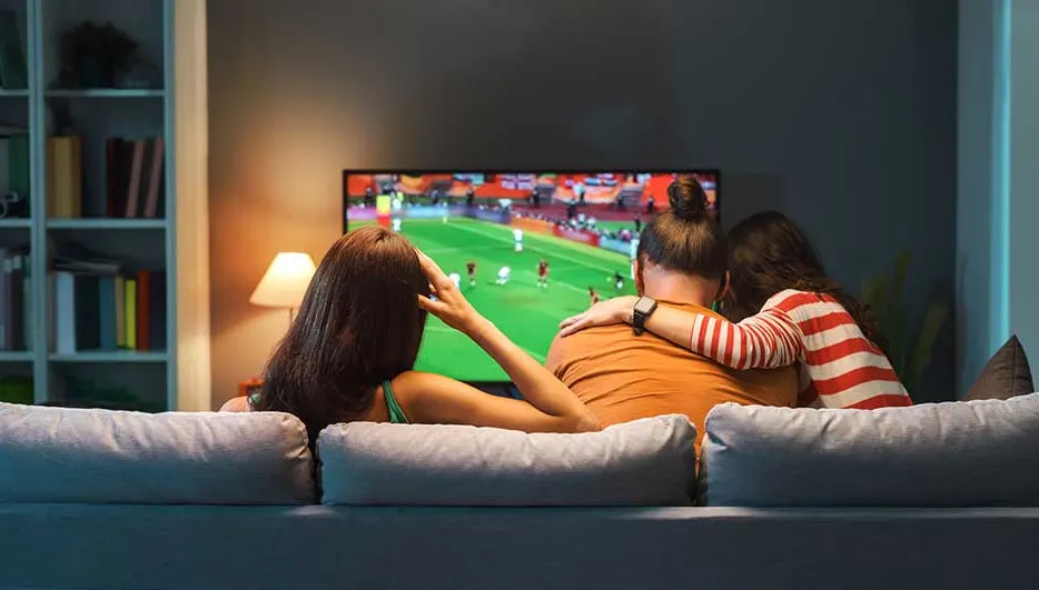 Trois personnes sont assises serrées sur un canapé et regardent un match de football à la télévision dans un salon chaleureux.