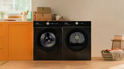 A laundry area with a stacked black washing machine and dryer. There's orange cabinetry, baskets, and folded towels nearby.