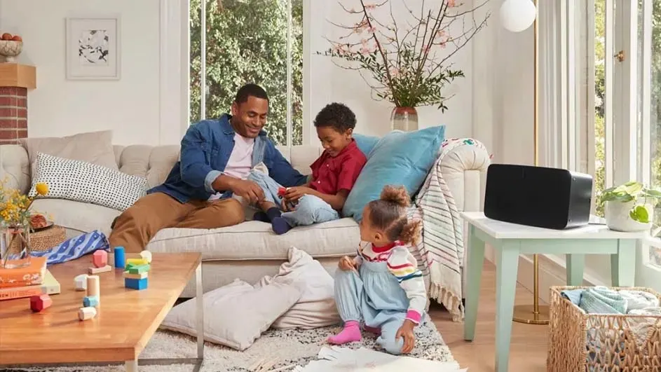 Dans un salon lumineux, un canapé beige avec des coussins colorés. Une table basse en bois présente des jouets. Un haut-parleur noir est sur une table d'appoint près d'une fenêtre.