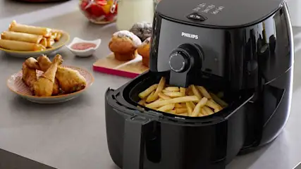 Een zwarte Philips airfryer met frieten in de mand, op een aanrecht met snacks en muffins.