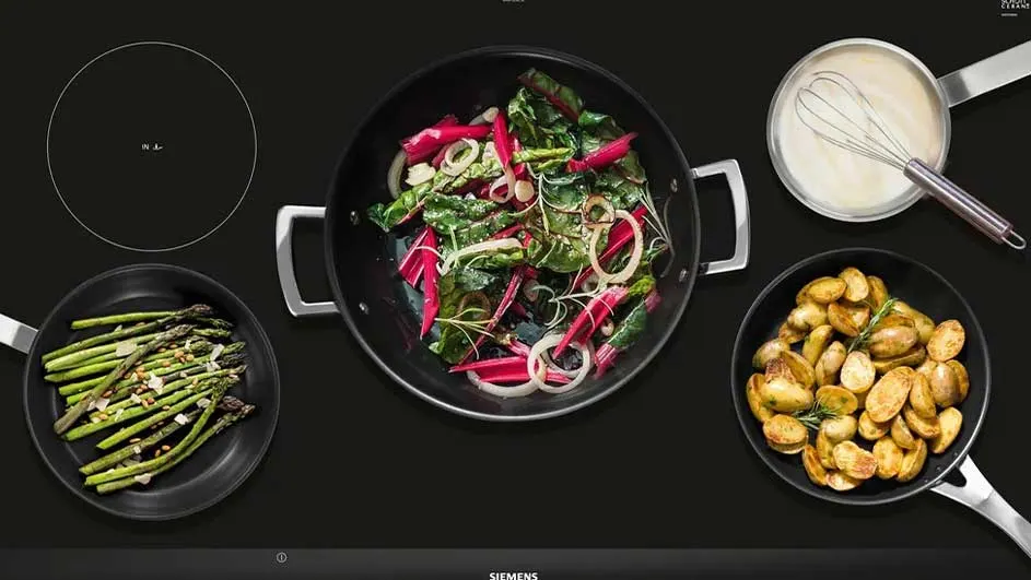 Een kookplaat met vier pannen: asperges, groentenmix, aardappelen en een saus met een garde.