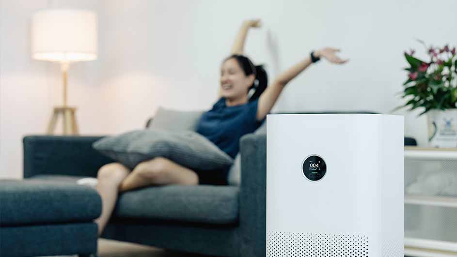 Un purificateur d'air blanc avec un écran rond est au premier plan. Un canapé gris et une lampe sont à l'arrière-plan.