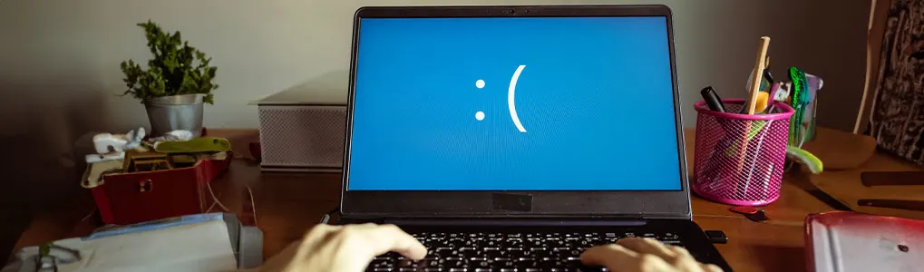 Un ordinateur portable affiche un écran bleu avec un visage triste sur un bureau encombré.
