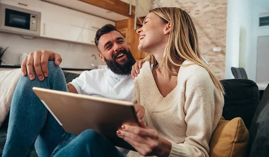 Un couple sur un canapé regarde une tablette. L'arrière-plan montre une cuisine blanche avec un four à micro-ondes.