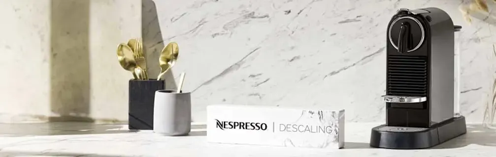 A Nespresso machine sits on a counter with a descaling kit, utensils in a holder, and a marble backdrop.