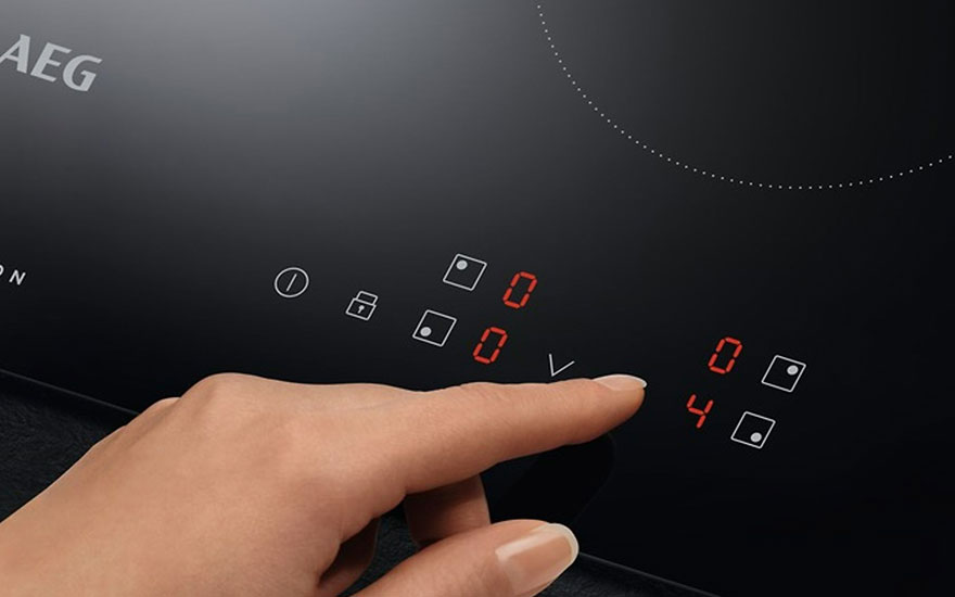 Een vinger bedient een zwarte AEG inductiekookplaat met rode digitale displays. De plaat heeft aan/uit knoppen en kookzones.