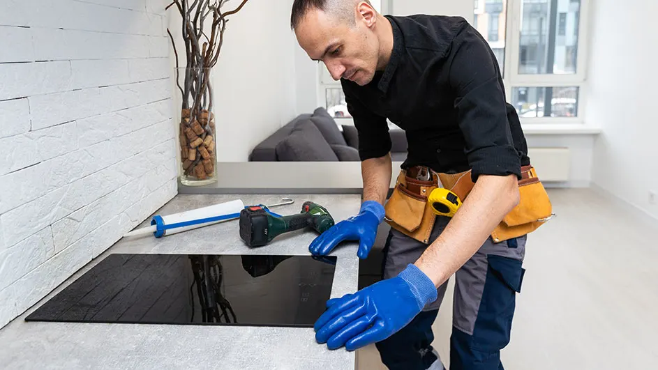 Un homme installe une plaque de cuisson noire sur un comptoir gris. Une perceuse et du mastic sont à côté.