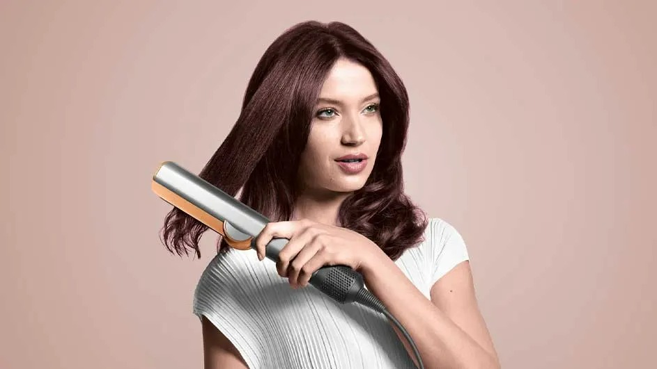 Une femme utilisant un fer à lisser argenté sur ses cheveux bruns devant un fond rose uni.