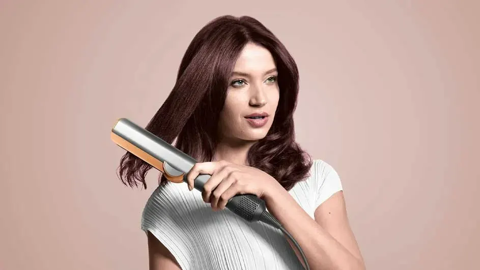 Une femme utilisant un fer à lisser argenté sur ses cheveux bruns devant un fond rose uni.