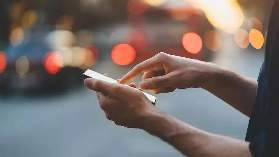 Hands hold a smartphone while one finger taps the screen; the background is a city street with blurred cars and warm bokeh lights at dusk.