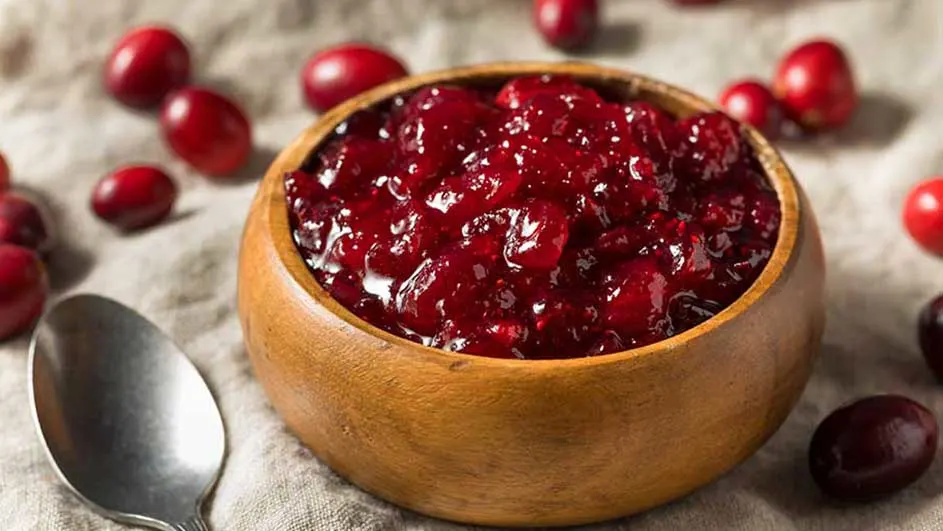 Een houten kom gevuld met rode veenbessencompote staat op een beige doek, met een zilveren lepel en losse veenbessen eromheen.