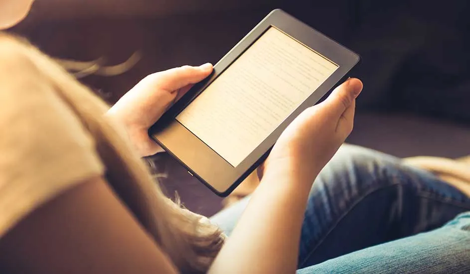 Een e-reader met een scherm met tekst wordt vastgehouden door twee handen tegen een achtergrond van blauwe jeans en een beige top.