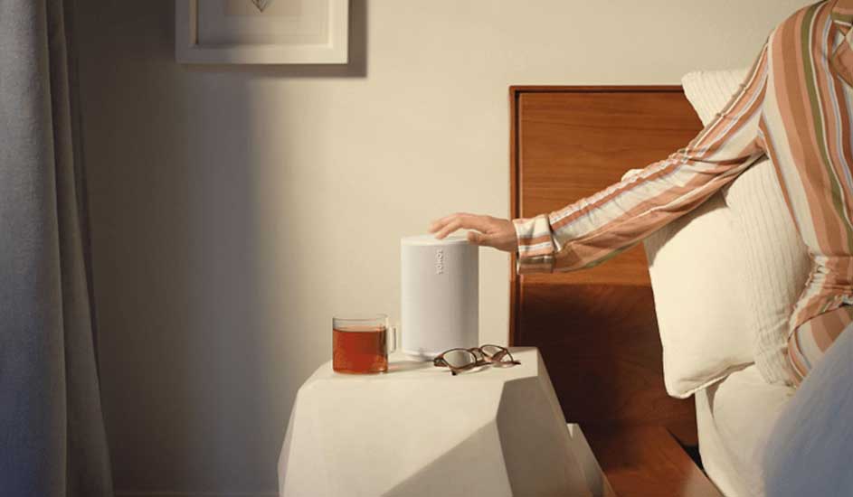 Een witte speaker staat op een wit nachtkastje naast een kop thee en een bril. Een hand raakt de speaker aan.