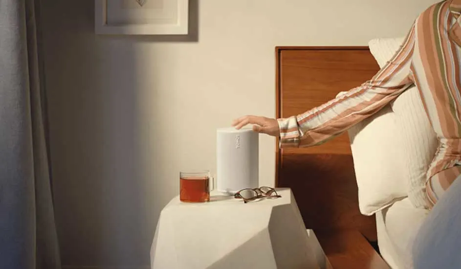 Een witte speaker staat op een wit nachtkastje naast een kop thee en een bril. Een hand raakt de speaker aan.