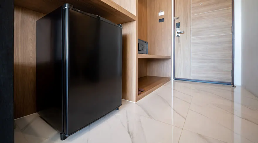 Vue d'une chambre d'hôtel avec un mini-frigo noir, des étagères en bois, un coffre-fort et un sol carrelé clair.