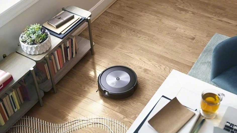 Een zwarte robotstofzuiger staat op een houten vloer in een kamer met boekenplanken en een witte tafel.