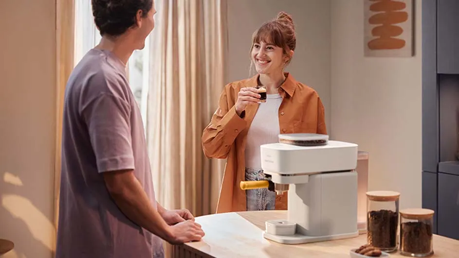 Une machine à café blanche est sur une table en bois, avec des bocaux de café à côté. Une femme tient une tasse.