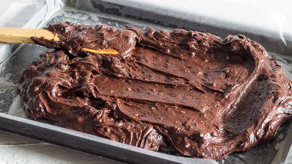 Een bakplaat met bruin browniebeslag wordt gladgestreken met een houten spatel op bakpapier.