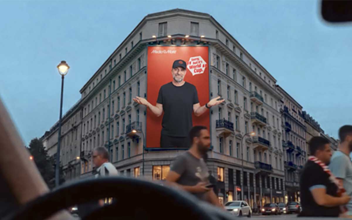 Un immeuble urbain affiche une immense publicité MediaMarkt avec un homme à l’écran, tandis que passants et voitures défilent.