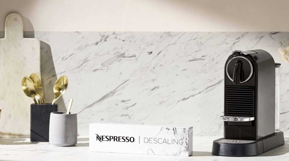 A kitchen scene featuring a black coffee machine, Nespresso descaling box, and gold utensils on a marble countertop.