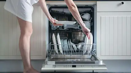 A person loads white plates into an open, stainless steel dishwasher in a white kitchen.