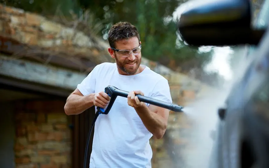 Un homme barbu avec lunettes de sécurité et t-shirt blanc lave sa voiture sombre avec un nettoyeur haute pression dans son jardin avec mur de briques