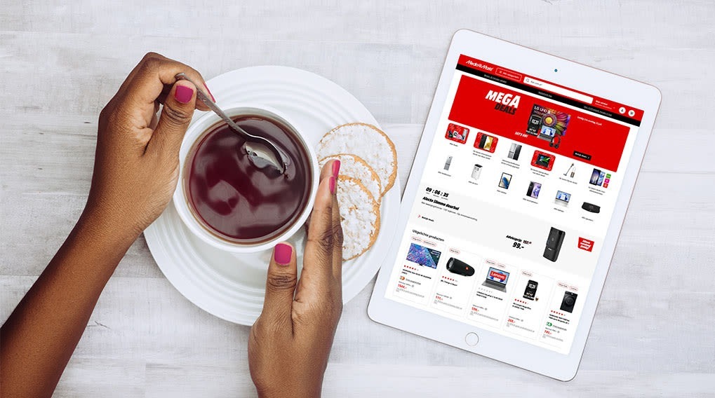 A tablet displays a website next to a plate with drink and cookies on a white wooden table.