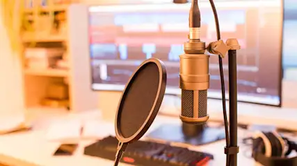Un microphone doré avec filtre anti-pop sur un bureau blanc, avec un ordinateur et un casque en arrière-plan.