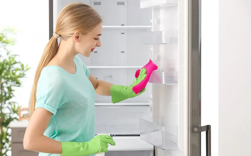 Une personne nettoie un réfrigérateur blanc avec des gants verts et un chiffon rose.