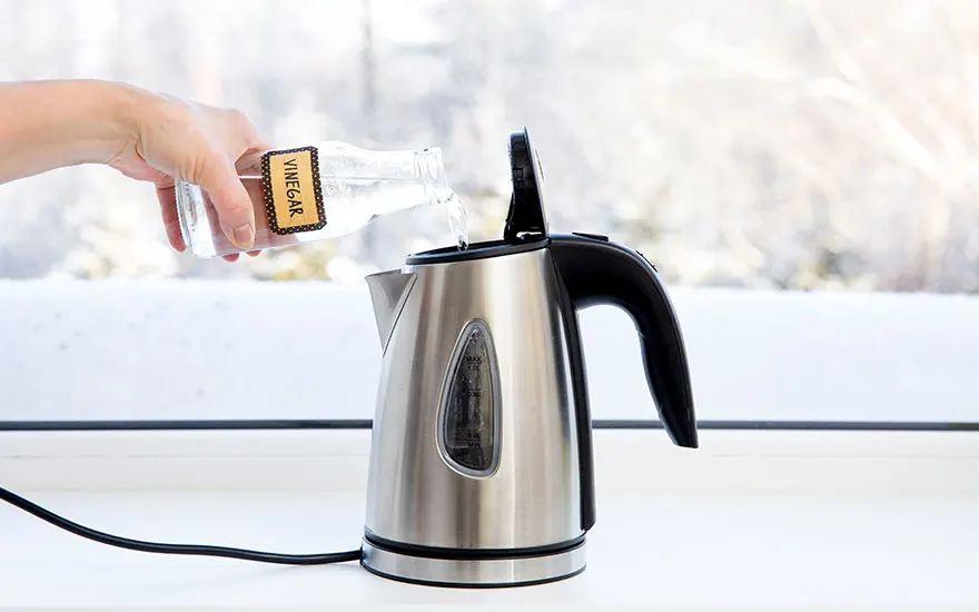 Une main verse du vinaigre d'une bouteille dans une bouilloire électrique en métal, sur un comptoir blanc.