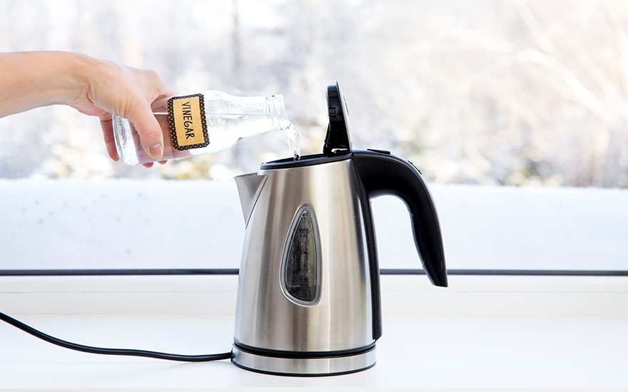 Een zilveren waterkoker staat op een wit aanrecht, met een fles azijn die erin wordt gegoten. Buiten een besneeuwd landschap.