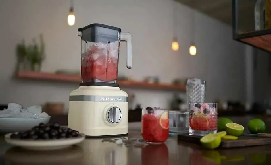 Un mixeur crème rempli d'une boisson rouge à côté de verres, de citrons verts et de myrtilles sur une table en bois.