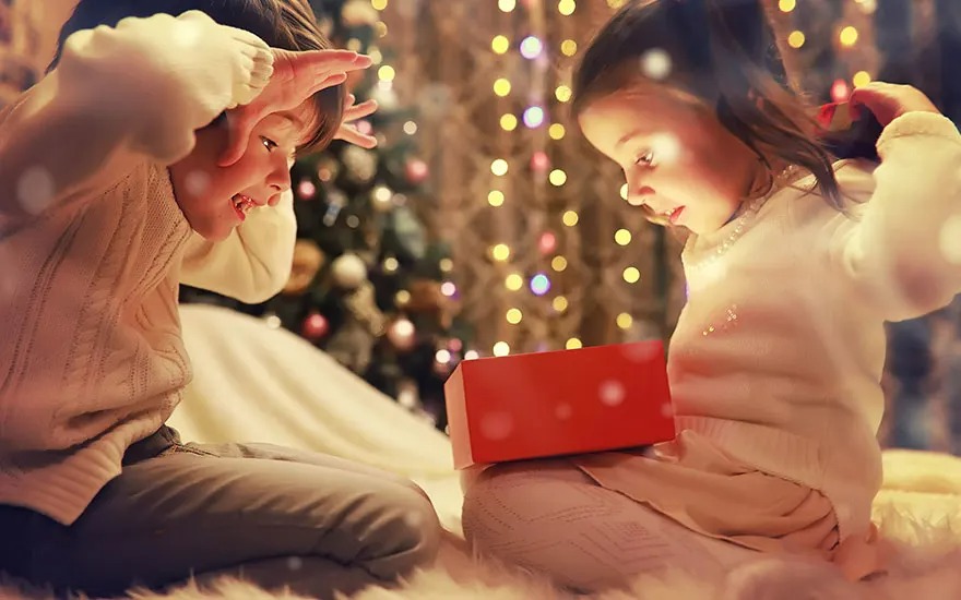Twee kinderen zitten op een zachte ondergrond, kijkend naar een rode doos. Op de achtergrond een verlichte kerstboom.