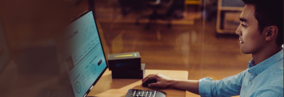 Een computer staat op een houten bureau met een zwart toetsenbord en muis. Achter de computer staat een zwarte doos.