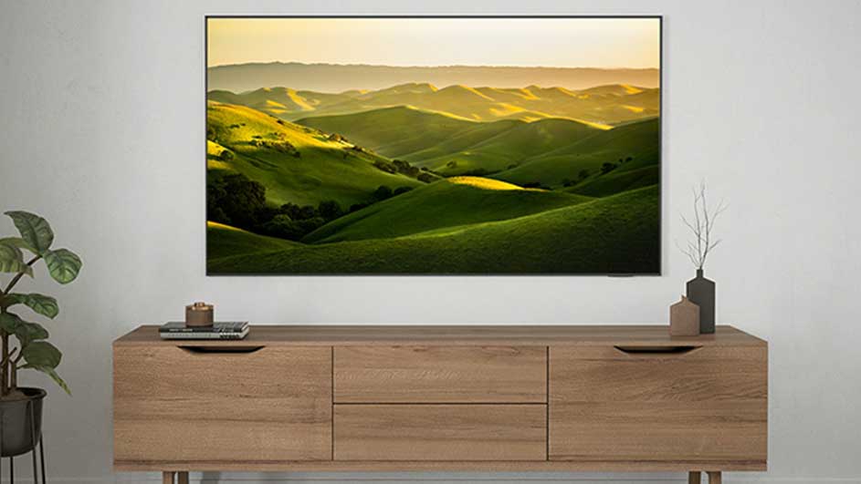 Een tv toont een groene heuvelrug. Onder de tv staat een houten dressoir met decoraties, met links een plant. De muur is wit.