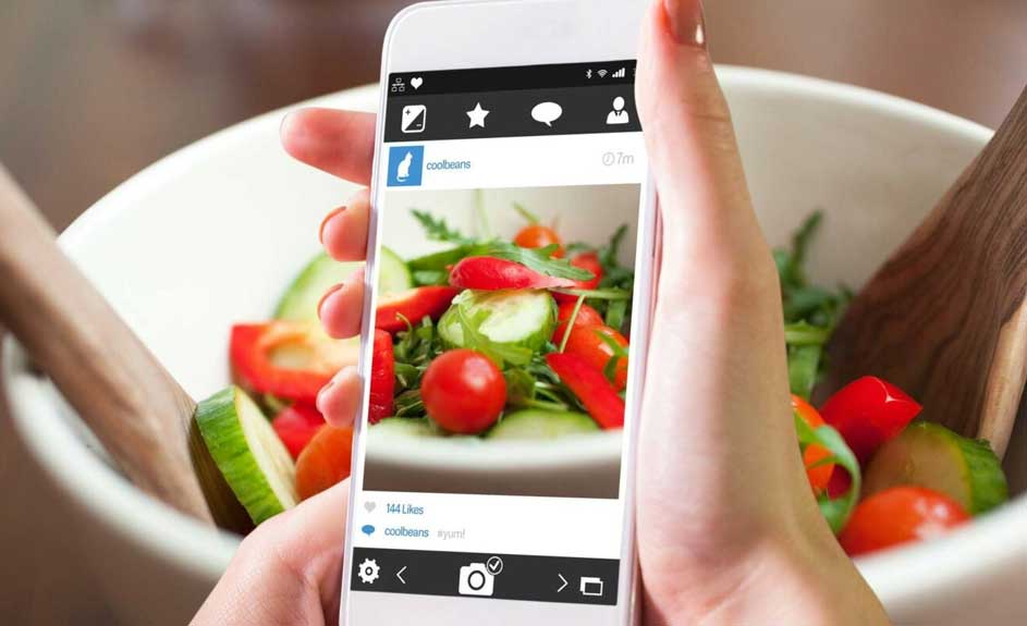 Een witte smartphone toont een saladefoto op een wit bord, voor een kom met salade en houten slacouvert.