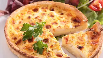 Une quiche dorée coupée, avec du persil frais. Salade verte et tomates en arrière-plan.
