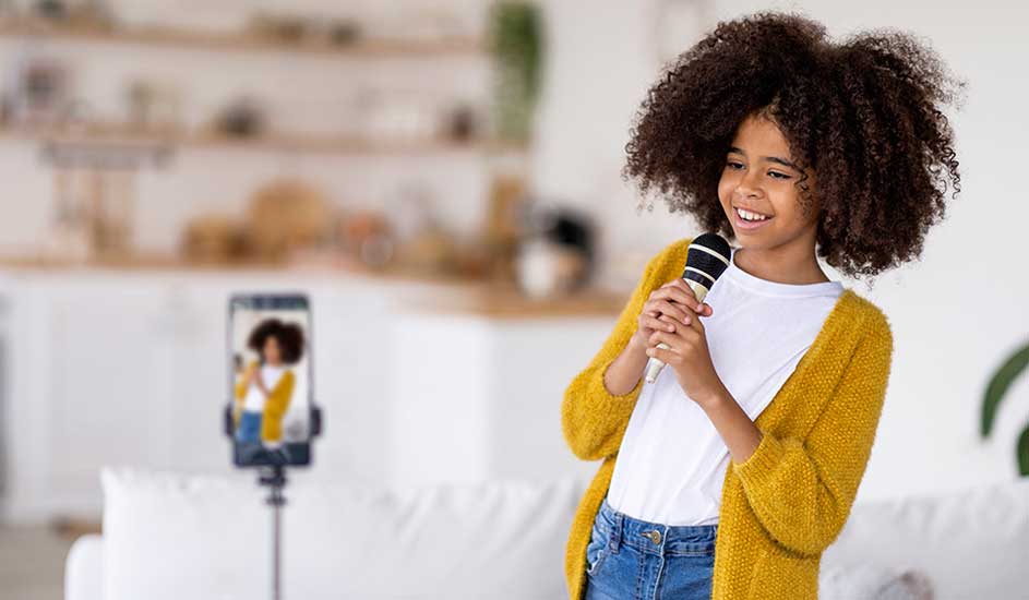 Une jeune fille chante dans un micro devant un smartphone sur un trépied. Elle porte un cardigan jaune et un jean.