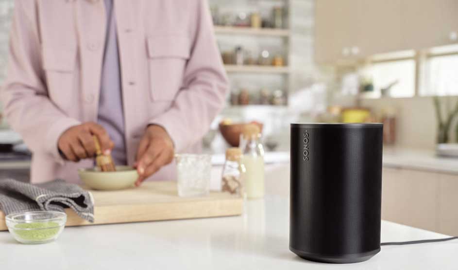 In een lichte keuken staat een zwarte speaker op het aanrecht, terwijl iemand op de achtergrond ingrediënten mengt in een kom.