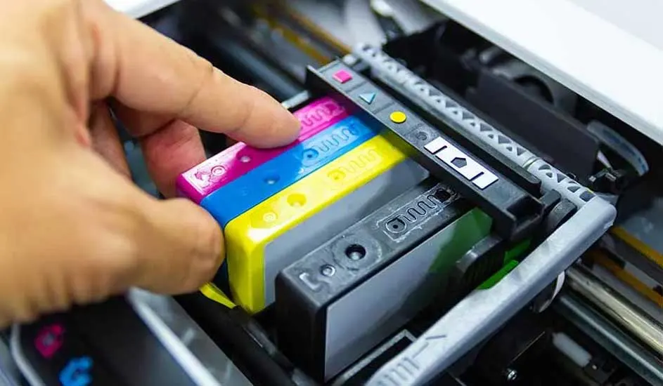 Een hand plaatst roze, blauwe en gele cartridges in een printer. De cartridges zijn naast een zwarte cartridge.