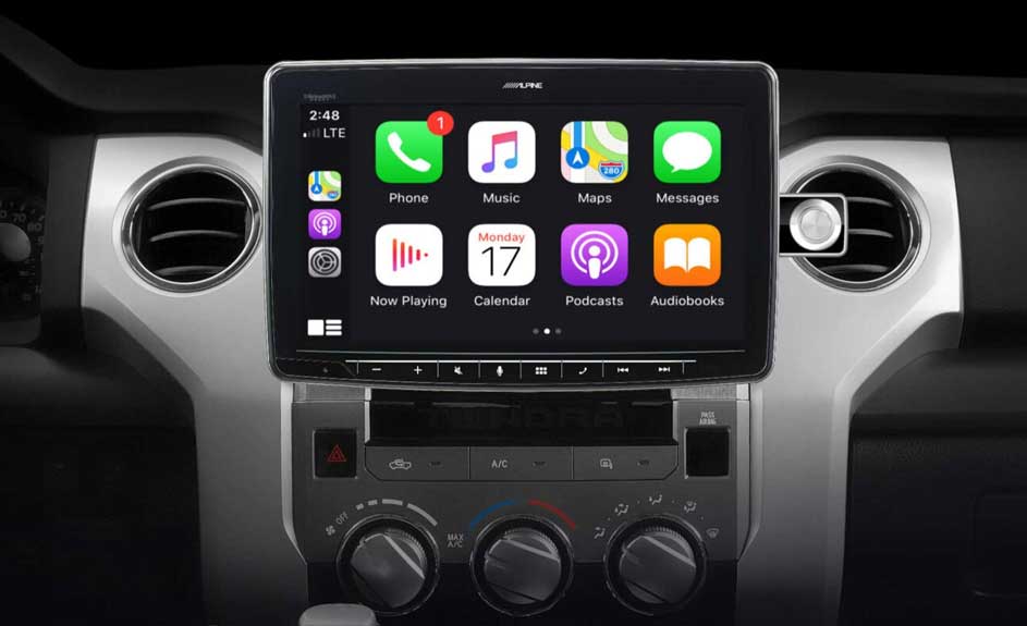 Auto-interieur met grijs dashboard. Centraal scherm toont apps: telefoon, muziek, kaarten, berichten, kalender, podcasts en audioboeken.
