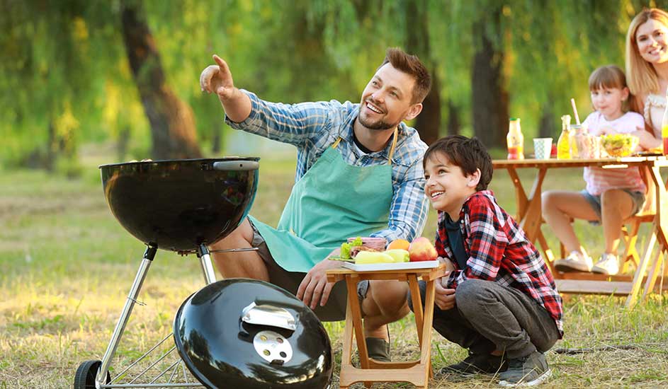 A man and a boy prepare a barbecue in a lush green garden, while a family eats and relaxes at a wooden table.