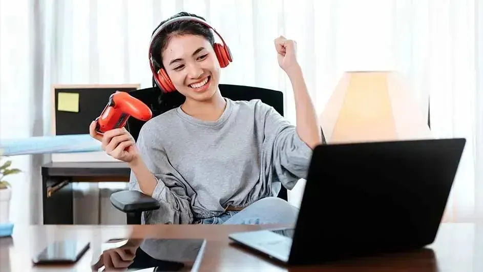 Young gamer wearing red headset cheers at laptop, raising controller while sitting at desk with lamp and phone.