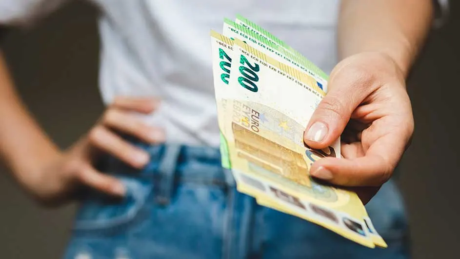 Een hand houdt biljetten van 200 euro vast tegen een achtergrond van een wit t-shirt en een blauwe jeans.