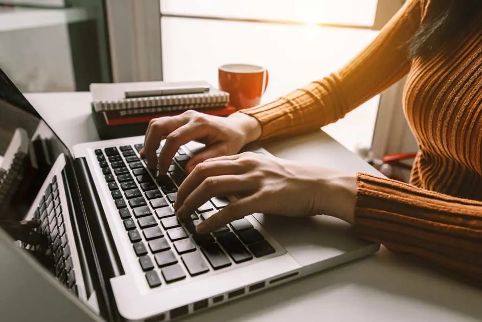 Une personne tape sur un ordinateur portable blanc sur un bureau blanc. Une tasse orange et des carnets sont à proximité.
