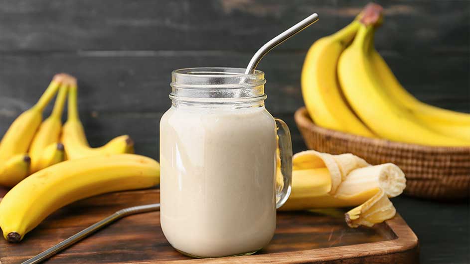 Un smoothie à la banane dans un bocal en verre avec une paille, entouré de bananes sur une planche à découper en bois.