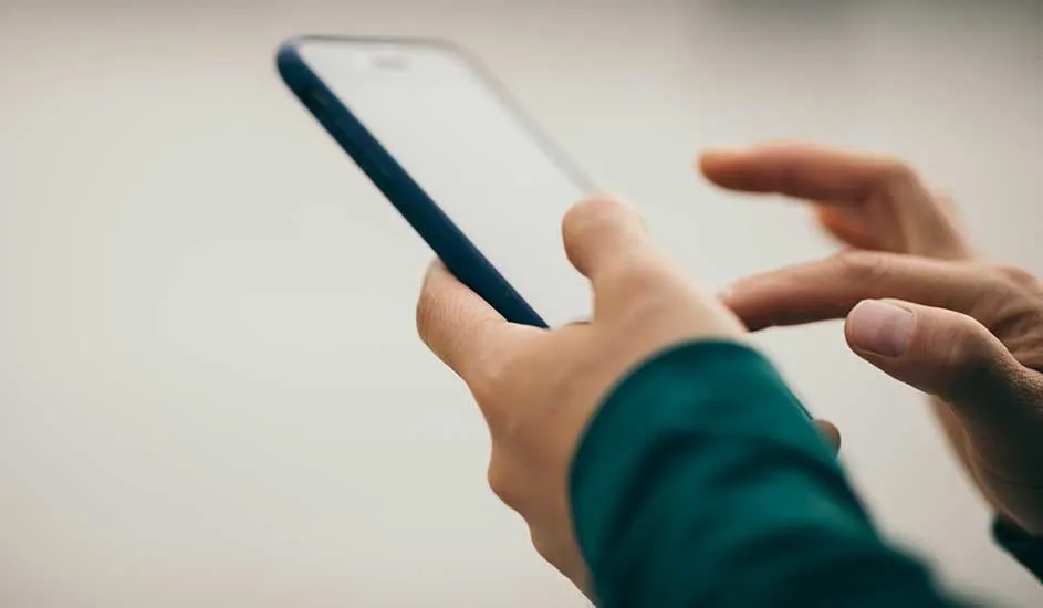 Een mobiele telefoon met een blauwe rand wordt tegen een vage, witte achtergrond vastgehouden. Een hand tikt op het scherm.