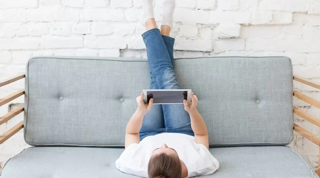 Une personne allongée sur un canapé gris, les jambes en l'air, tenant une tablette devant un mur de briques blanches.