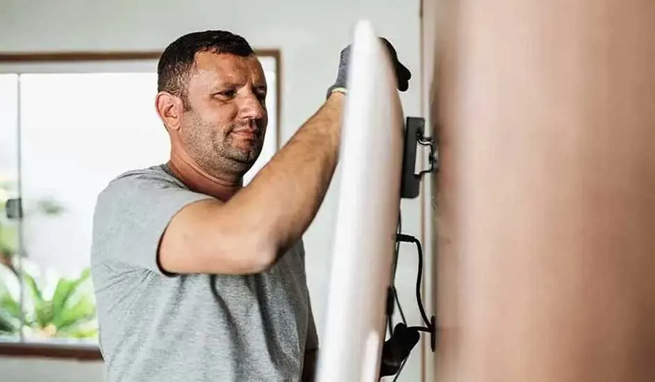 Someone is mounting a flat-screen TV on the wall in a bright room, wearing gloves for safety. Brand is not visible.
