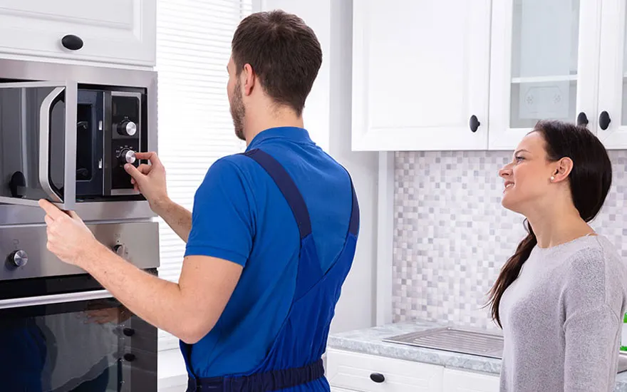 Een technicus in blauwe overall controleert een magnetron in een witte keuken. Aanrecht en tegels zijn zichtbaar.